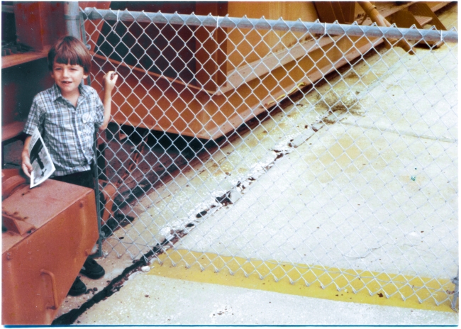 Kai MacLaren stands next to the lower drive and support portion of the Redstone Gantry at Complex 26, Cape Canaveral Air Force Station, Florida. Visible through the chain-link fencing in the shadowed area to his right, the rails that this gantry traveled upon can be seen. Photograph by James MacLaren.